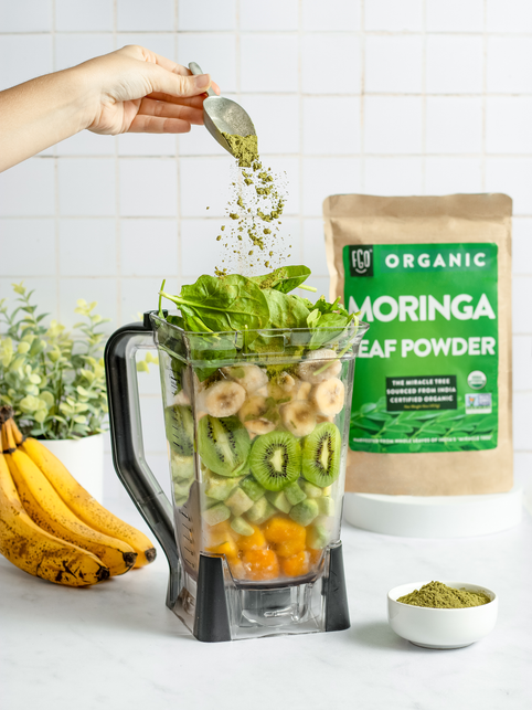 Moringa leaf powder sprinkled into a blender full of fresh greens and fruits.