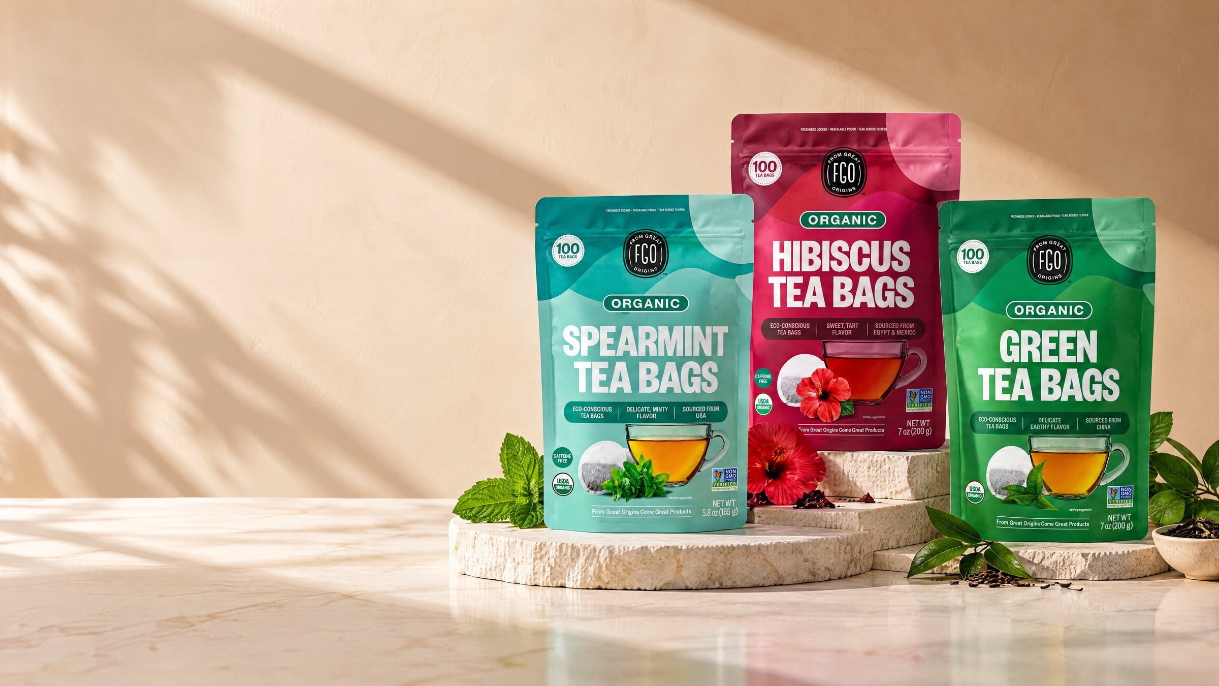 Spearmint, Hibiscus and Green Tea Bags pouches on stone risers in warm afternoon light, with fresh mint, hibiscus flowers and tea leaves beside them