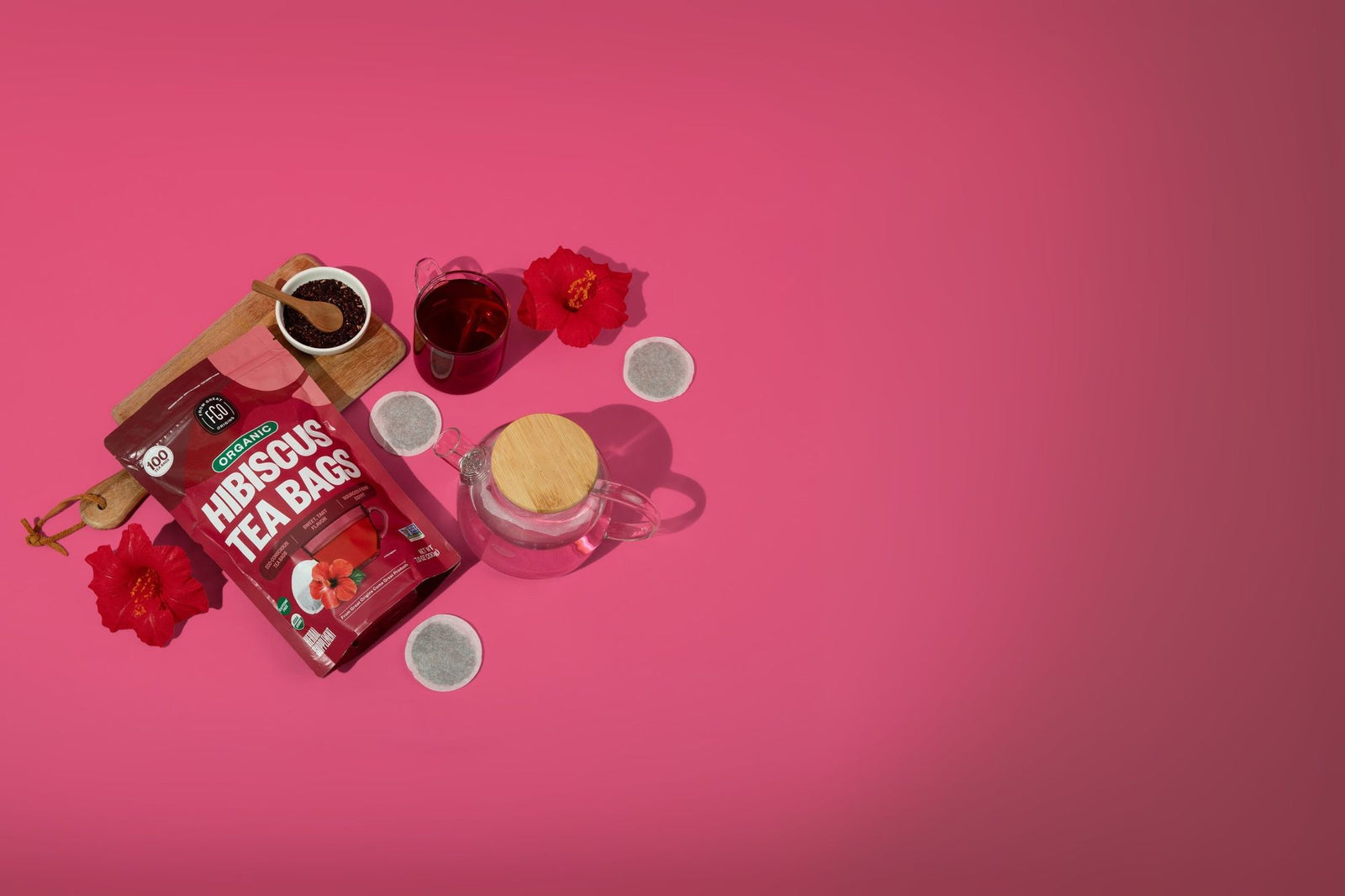 A pink flat lay with a Hibiscus Tea Bags pack, a bowl of dried hibiscus and a glass of hibiscus tea